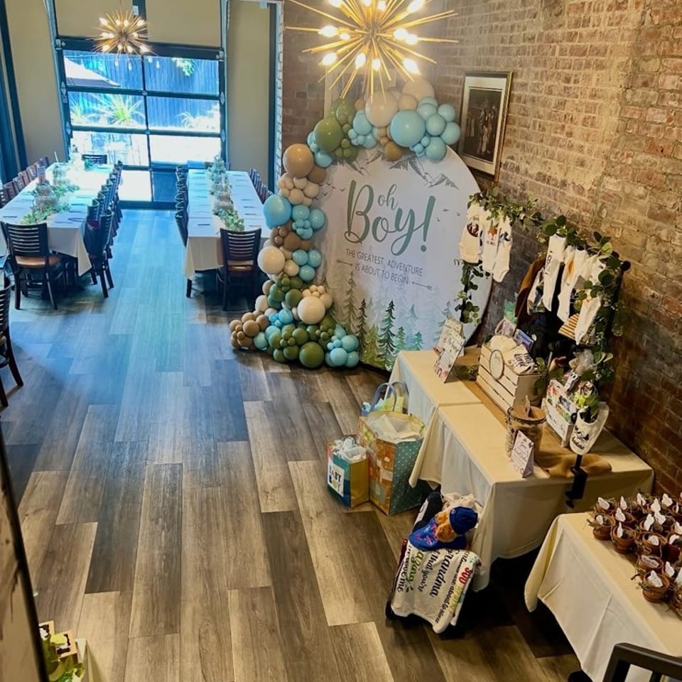A baby shower setup with an "Oh Boy!" sign, balloons, gifts, and dining tables in a restaurant.
